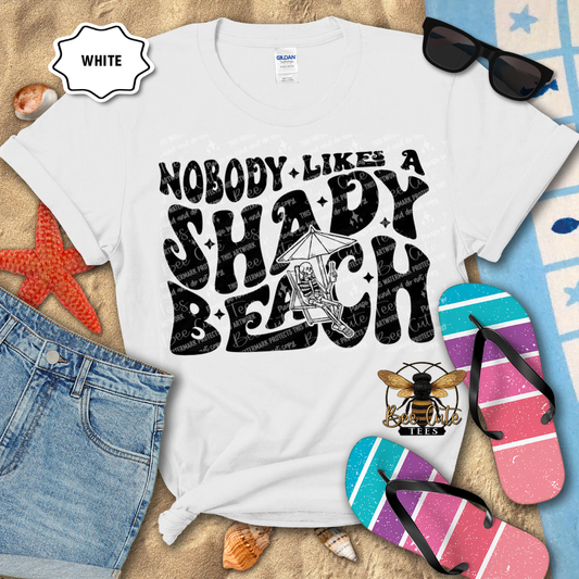 White t-shirt with 'Nobody Likes a Shady Beach' text, sunglasses, and flip-flops on a beach-themed background.