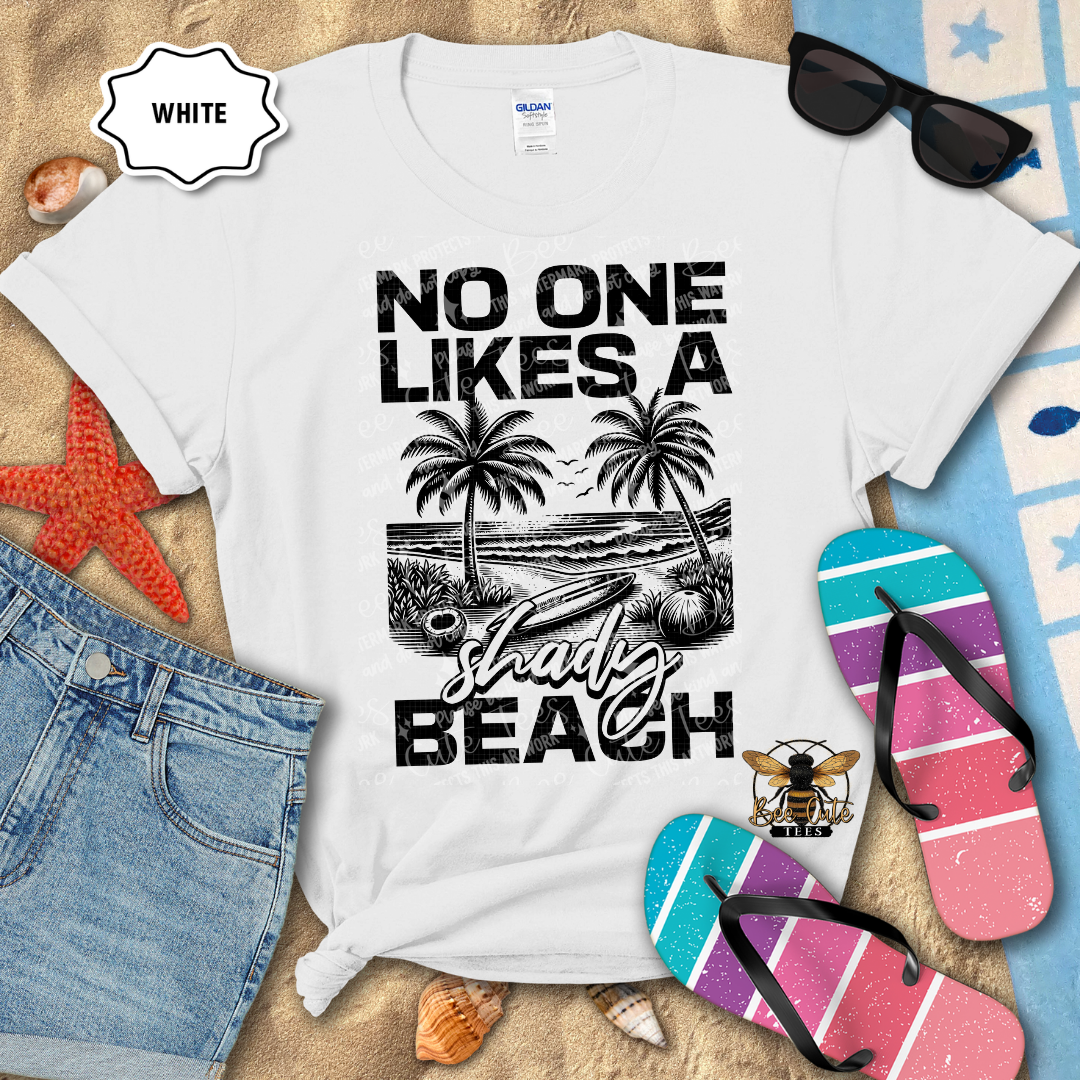 White t-shirt with beach-themed design and text, surrounded by flip-flops and sunglasses on a sandy background.