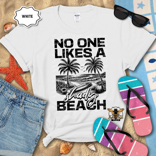White t-shirt with beach-themed design and text, surrounded by flip-flops and sunglasses on a sandy background.