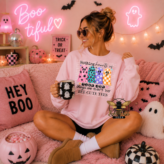 A woman sits cross-legged on a pink couch wearing a light pink graphic sweatshirt with a Halloween-themed design. The sweatshirt features a colorful ghost pattern and the text 'nothing hearts us when we don't buy BEE CUTE TEES'.