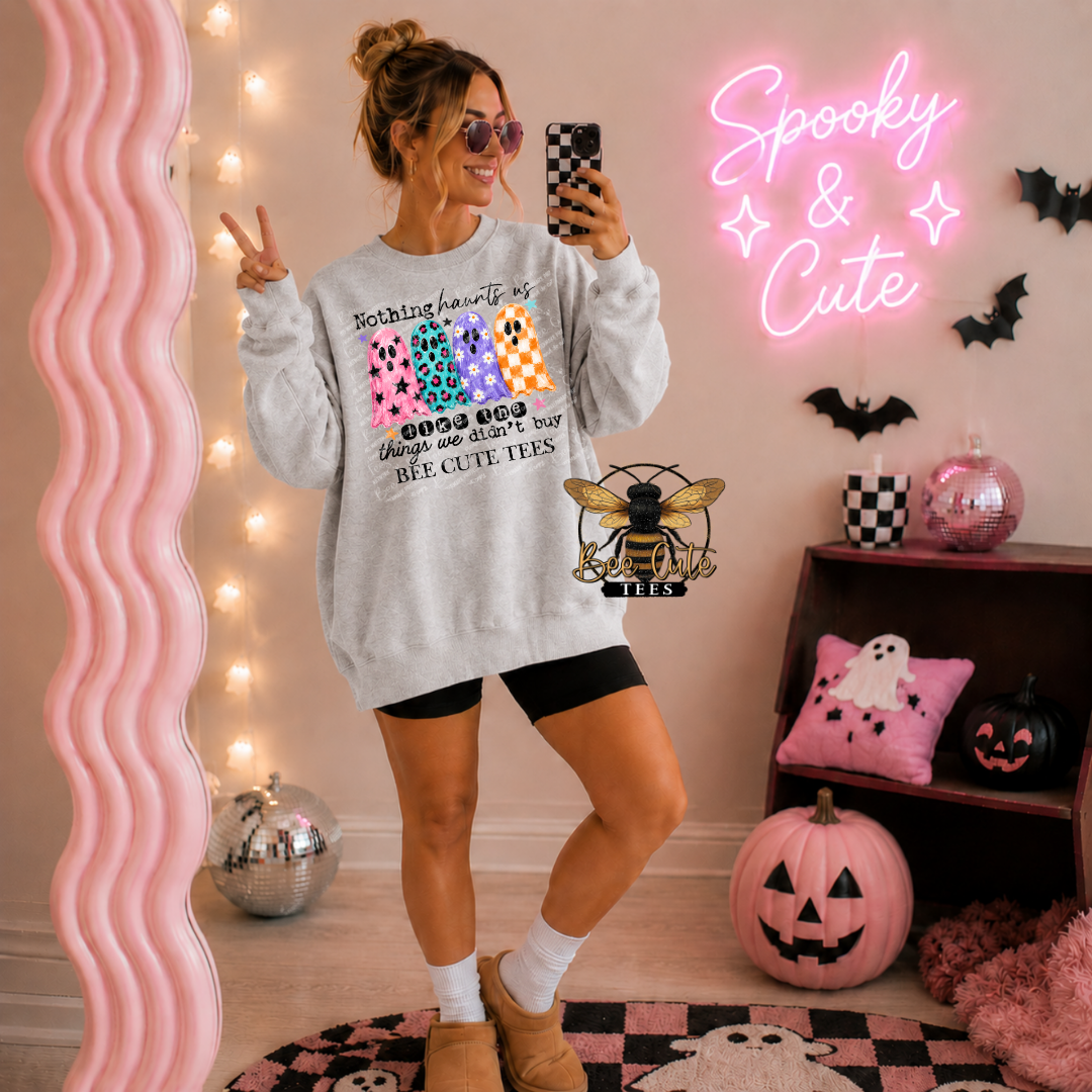 A woman takes a mirror selfie wearing a light gray crewneck sweatshirt with a colorful 'Spooky' graphic, black bike shorts, and tan boots in a decorated Halloween room with a pink neon sign and pumpkins.