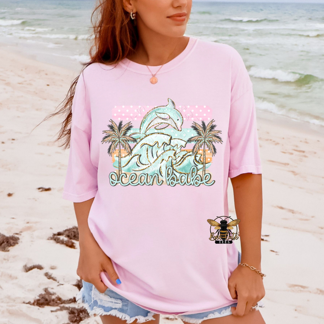 Person wearing a pink t-shirt with a dolphin and palm tree design on a beach.