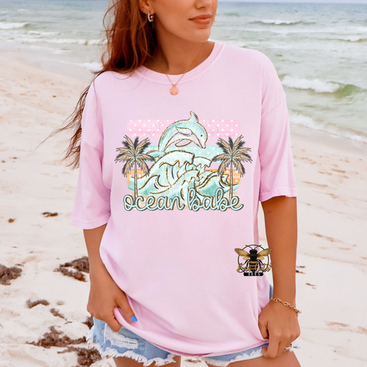 Person wearing a pink t-shirt with a dolphin and palm tree design on a beach.
