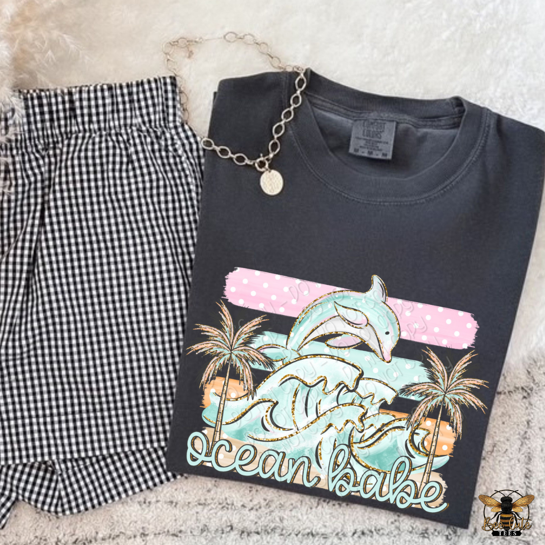 Black t-shirt with a dolphin graphic and 'Ocean Babe' text, paired with checkered pants on a textured surface.