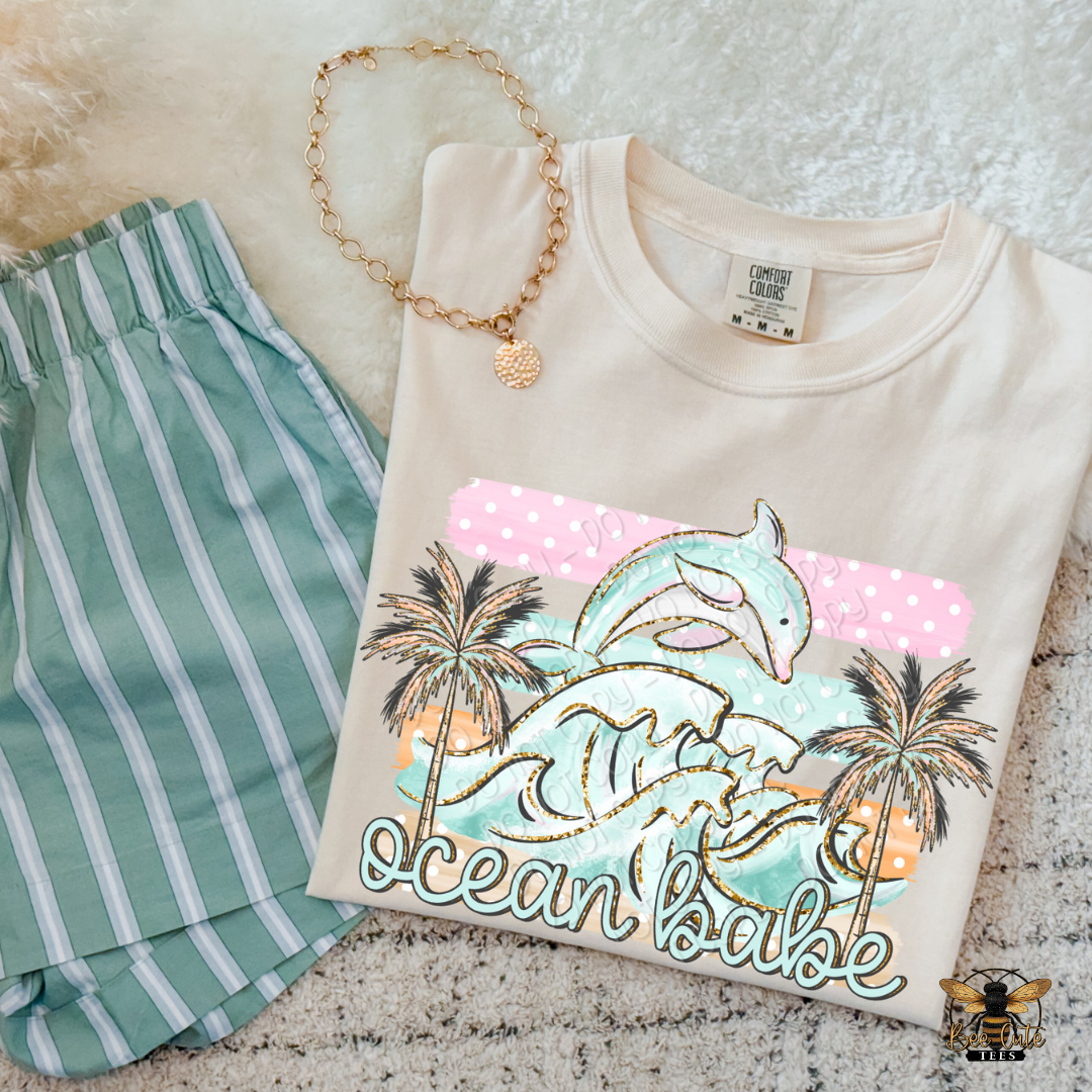 Beige sweatshirt with 'ocean babe' design, green striped pants, and gold necklace on textured surface