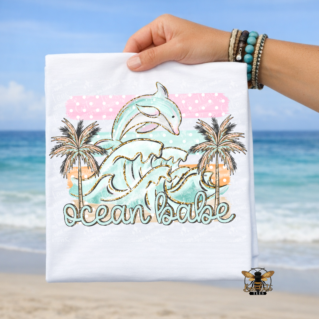 Hand holding a white t-shirt with a dolphin and palm tree design against a beach background