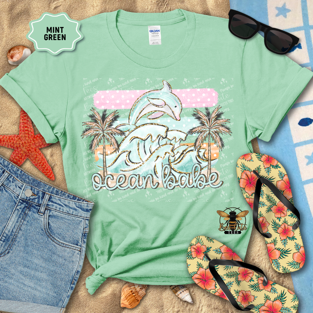 Mint green t-shirt with 'ocean babe' design, sunglasses, flip-flops, and beach accessories on a sandy background.