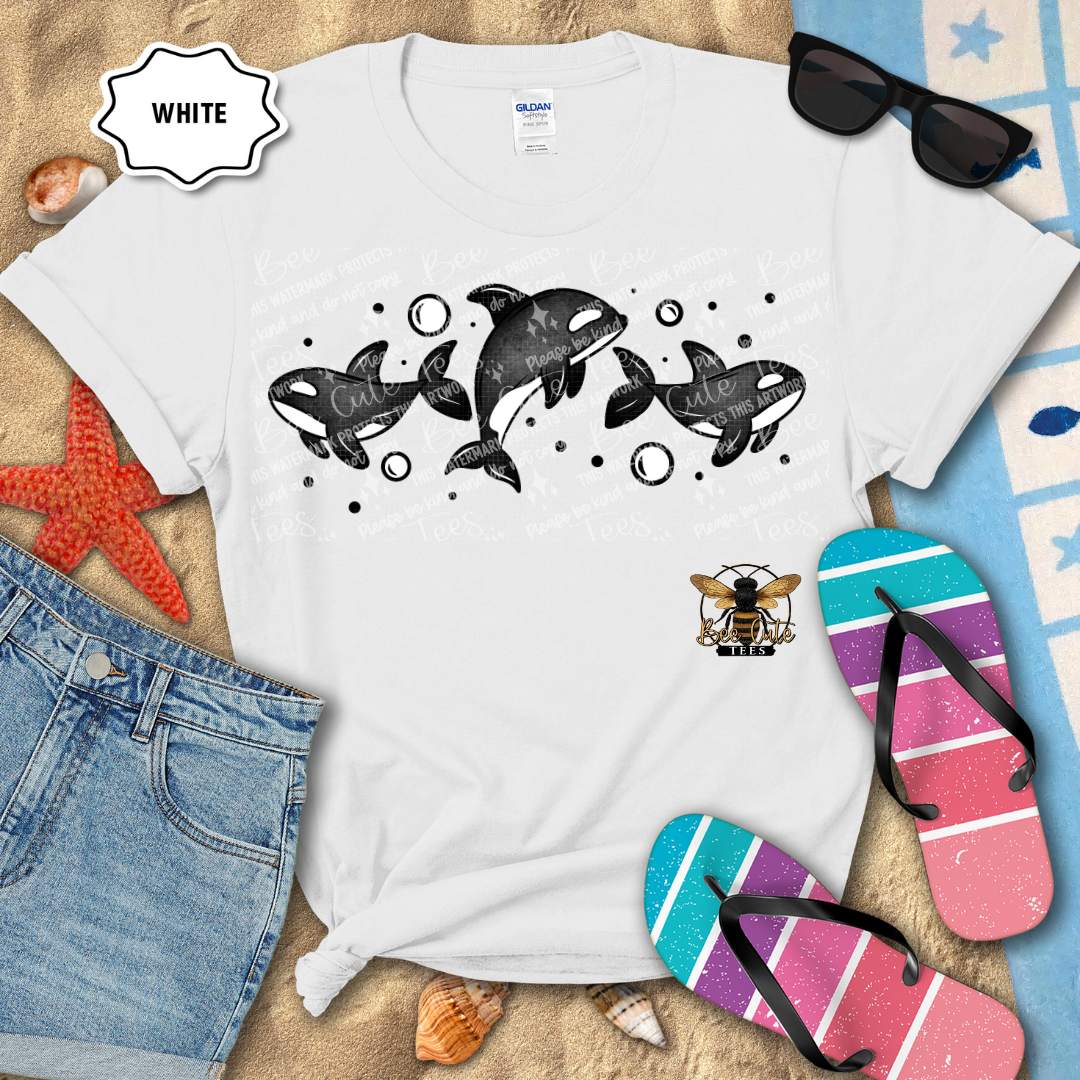 White t-shirt with orca design on a sandy surface with beach accessories.