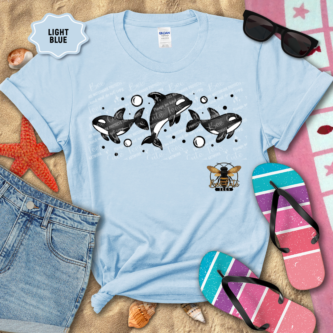 Light blue t-shirt with orca whale design on a sandy surface with beach accessories.