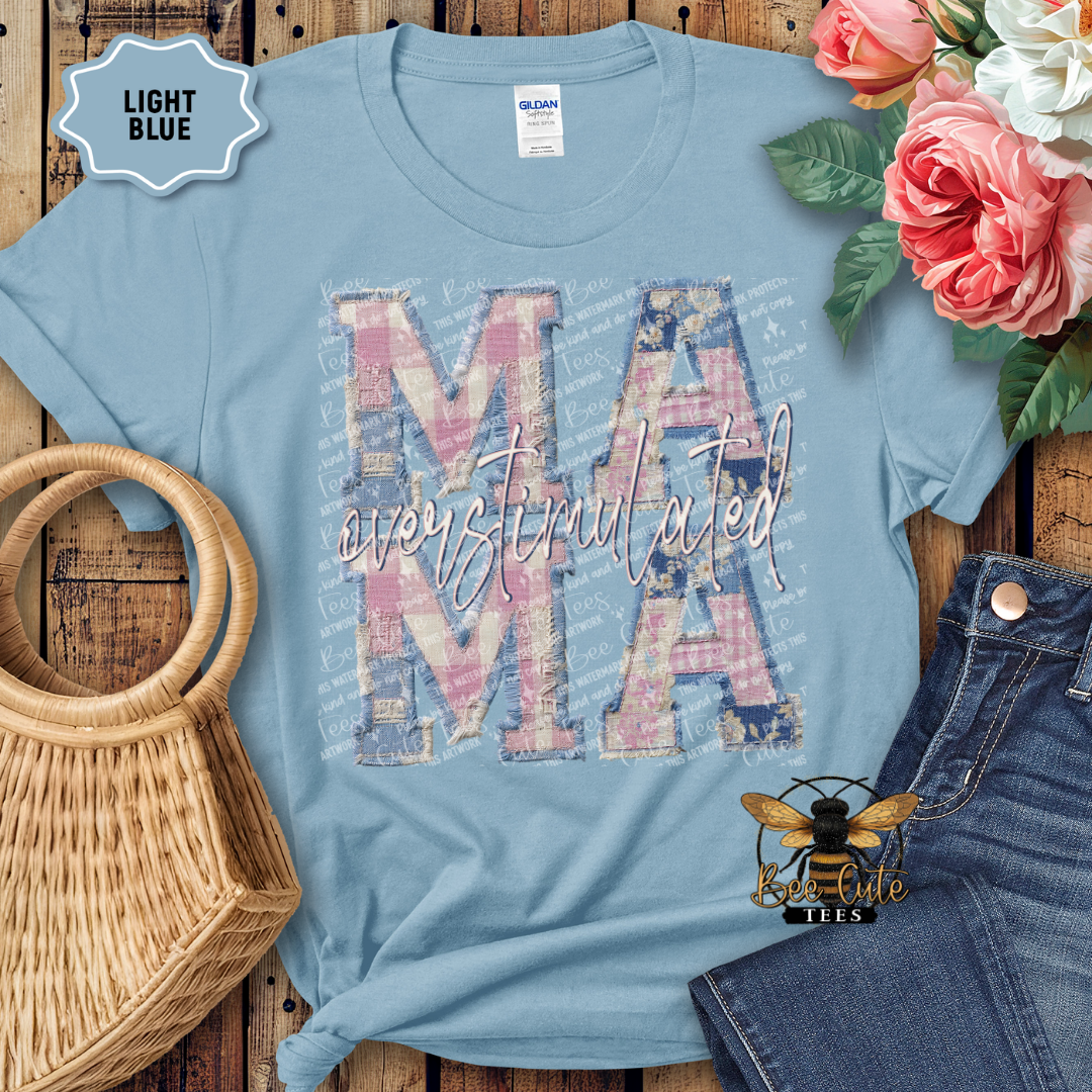 Light blue t-shirt with 'overstimulated MAMA' design, surrounded by flowers and denim jeans on a wooden surface.