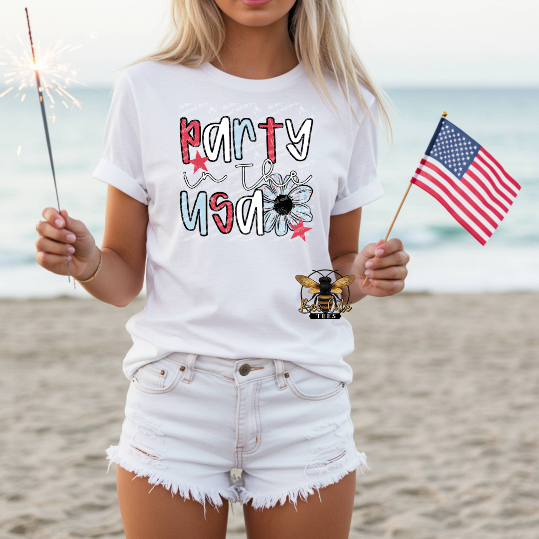 Person wearing a 'Party USA' t-shirt on a beach with an American flag and sparkler.