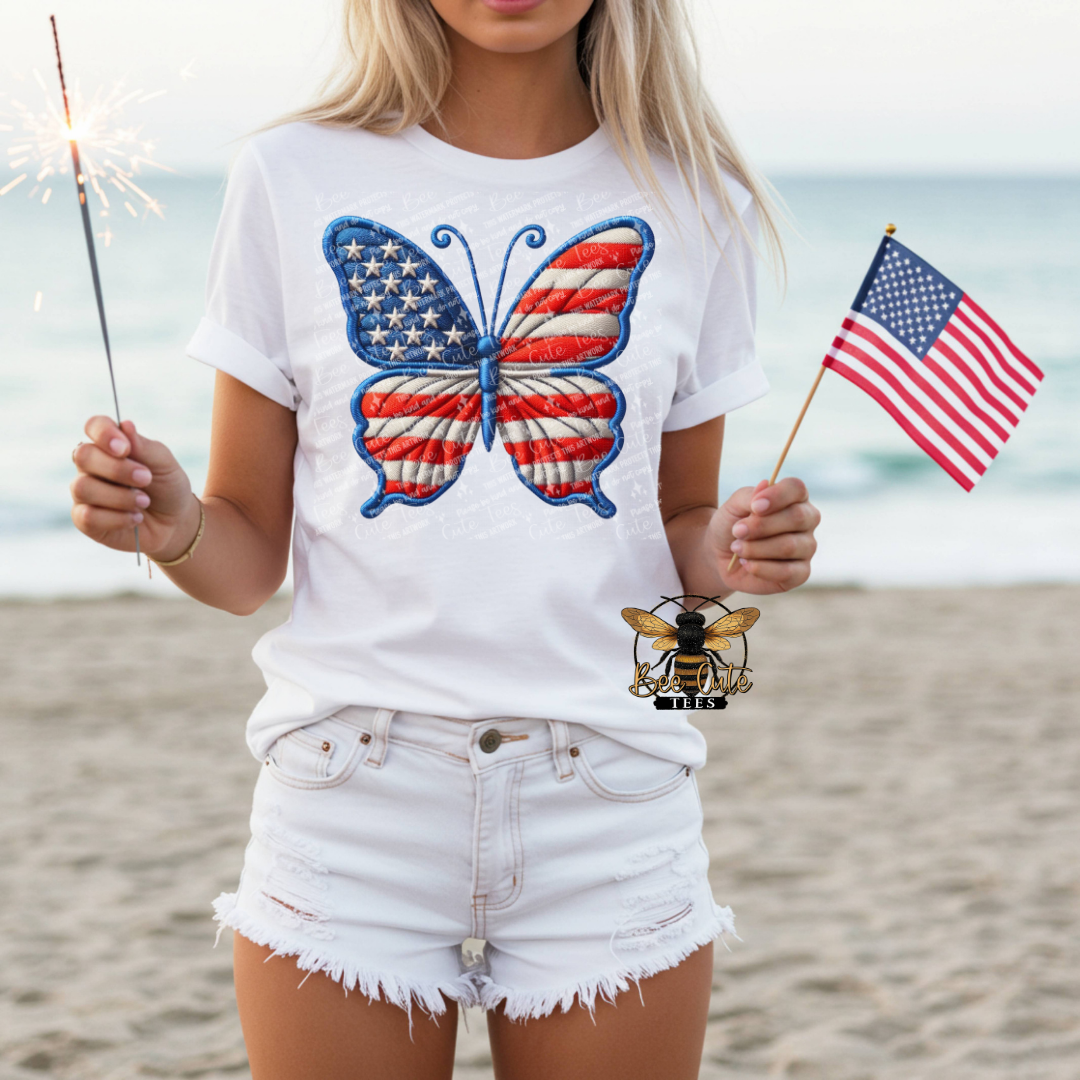 Person on a beach wearing a white t-shirt with a butterfly design holding an American flag and a sparkler.