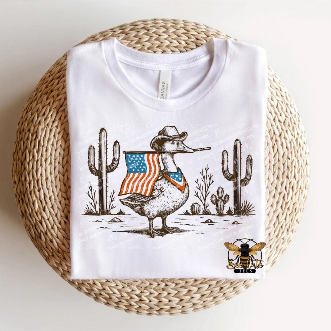 White t-shirt with a graphic of a duck wearing a cowboy hat and American flag, placed on a woven surface.