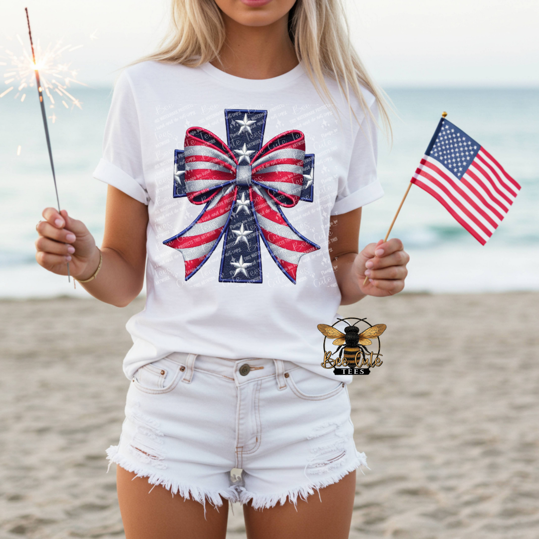 Person on a beach wearing a white t-shirt with a red, white, and blue cross design, holding an American flag and a sparkler.