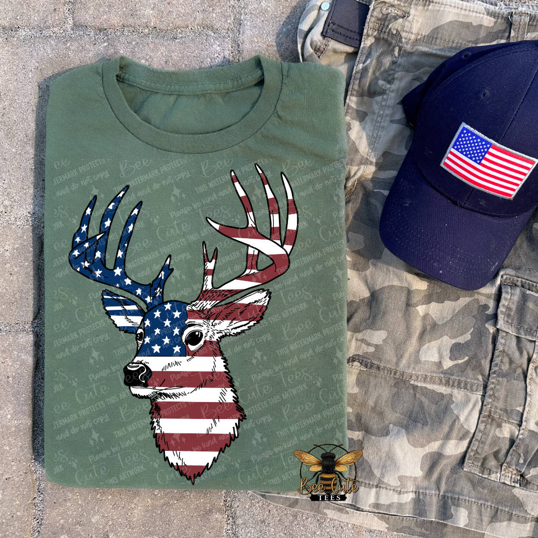Green t-shirt with a deer head design featuring American flags, next to a blue cap with an American flag patch on a textured surface.