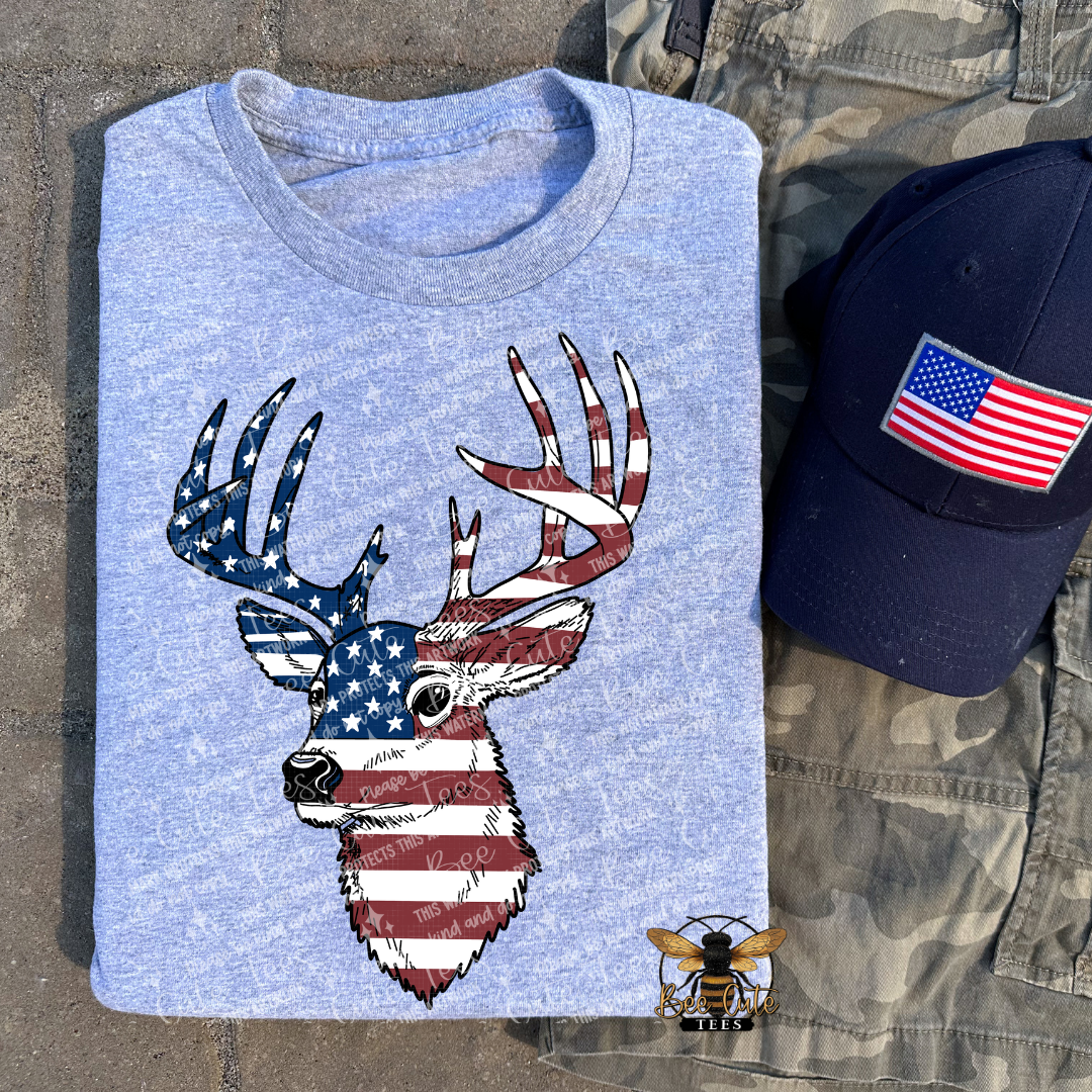 Grey t-shirt with a deer head design featuring the American flag, next to a cap with an American flag patch on a stone surface.