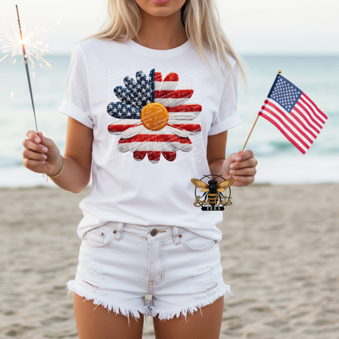 Person wearing a white t-shirt with a colorful design, holding an American flag and a sparkler on a beach.
