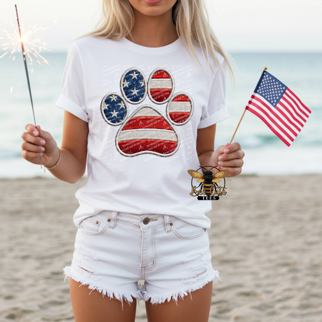 Person on a beach wearing a white t-shirt with a paw print design and holding an American flag.