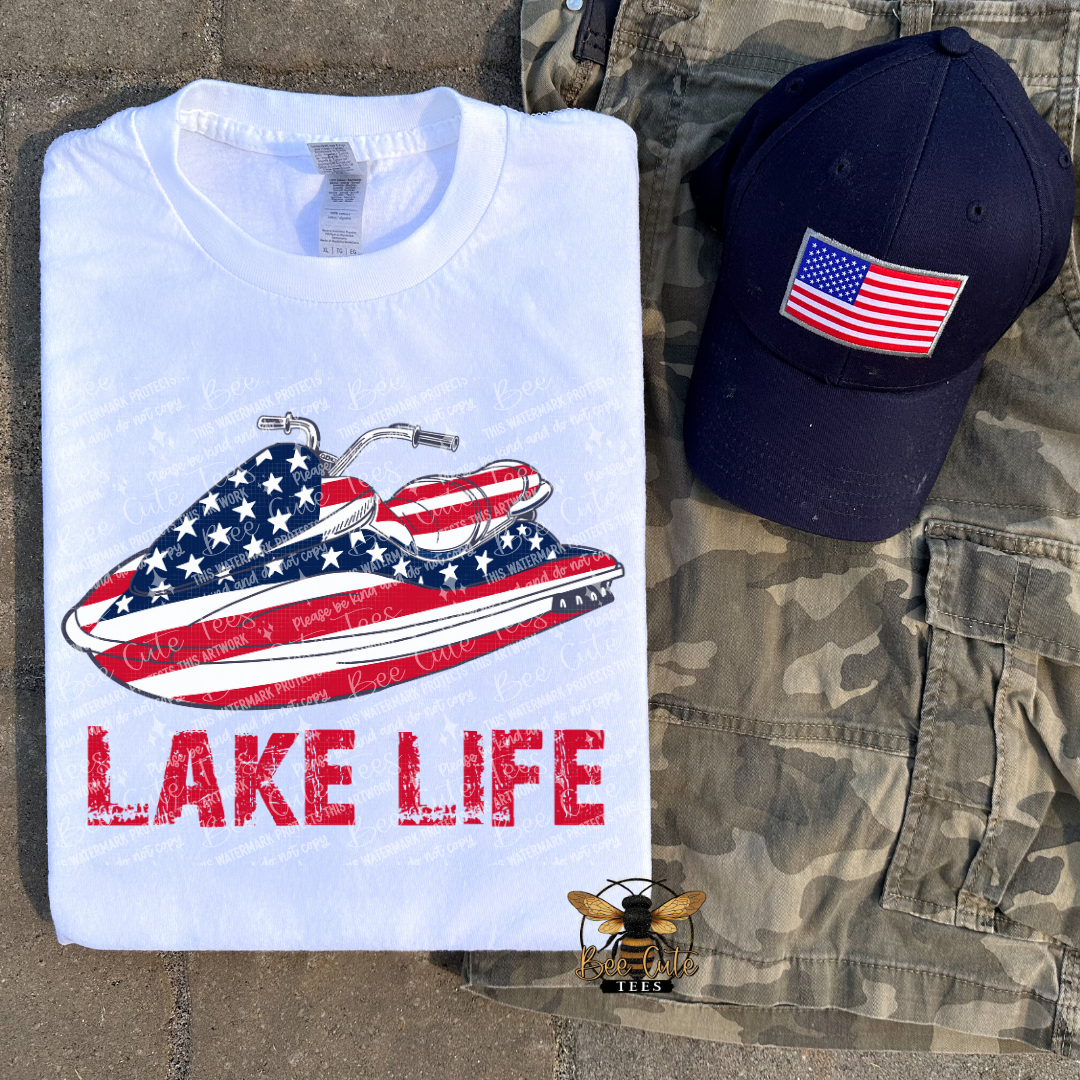 White t-shirt with American flag jet ski design and 'Lake Life' text, navy cap with American flag, and camouflage shorts on a stone surface.
