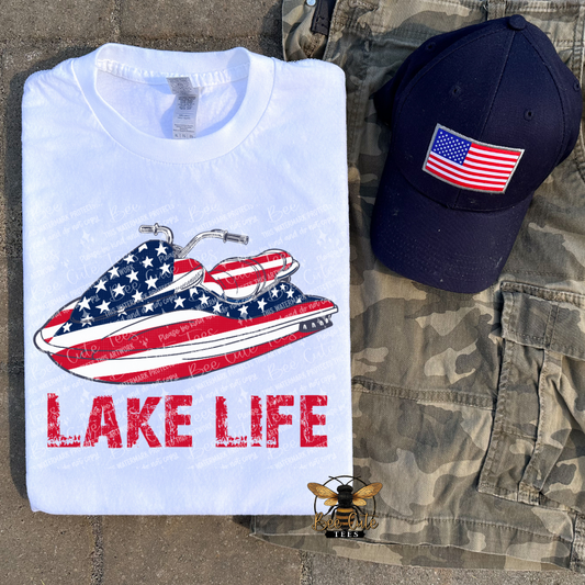 White t-shirt with American flag jet ski design and 'Lake Life' text, navy cap with American flag, and camouflage shorts on a stone surface.