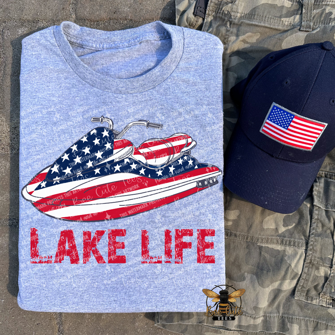 Grey t-shirt with American flag jet ski design and 'Lake Life' text, next to a navy cap with an American flag patch.