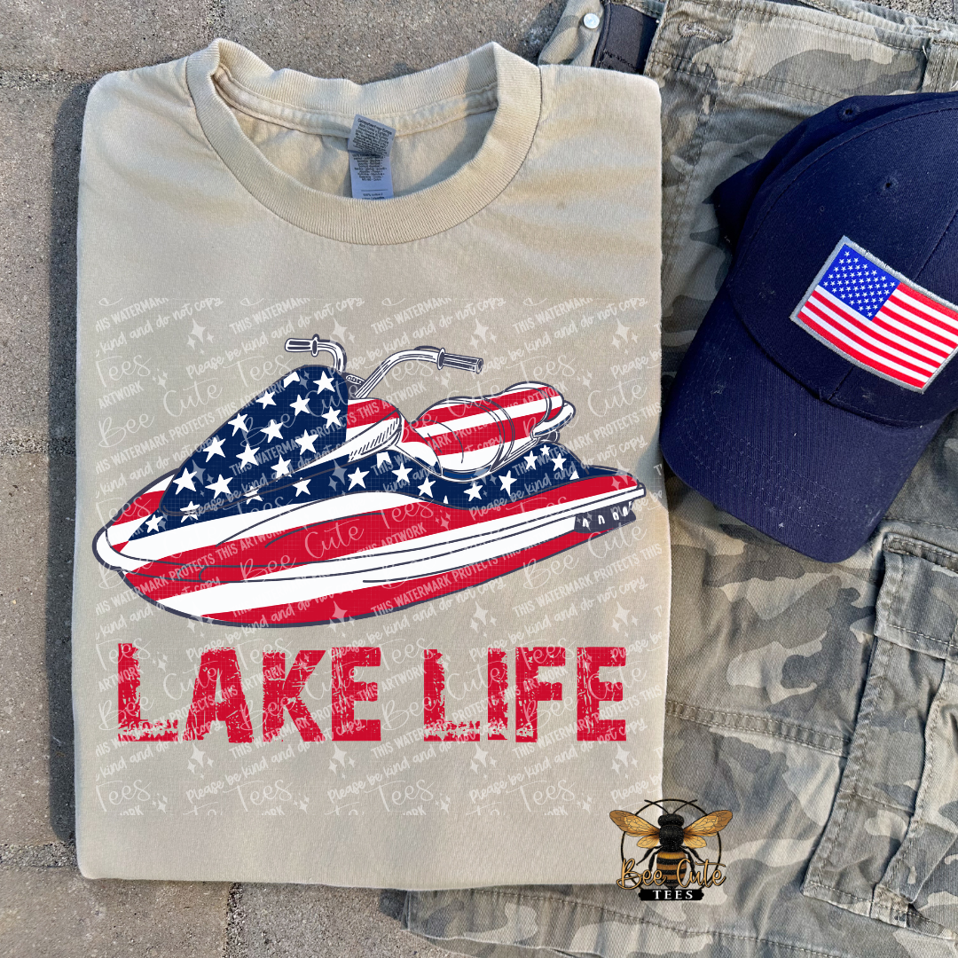 Folded t-shirt with American flag jet ski design and 'Lake Life' text, next to a cap with an American flag patch.
