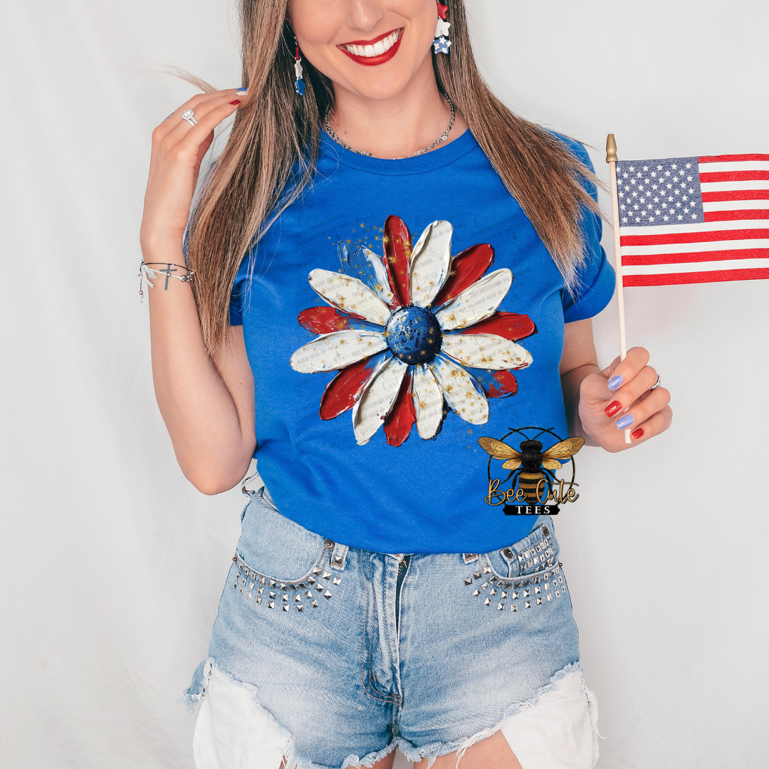 Woman wearing a royal blue graphic tee featuring a distressed patriotic daisy design with red, white, and blue petals and a navy blue center. Styled with light-wash denim shorts, patriotic earrings, and holding a small American flag, creating a festive Americana look perfect for summer celebrations.
