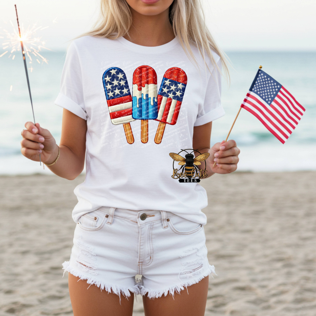 Person wearing a white t-shirt with patriotic popsicle design on a beach.