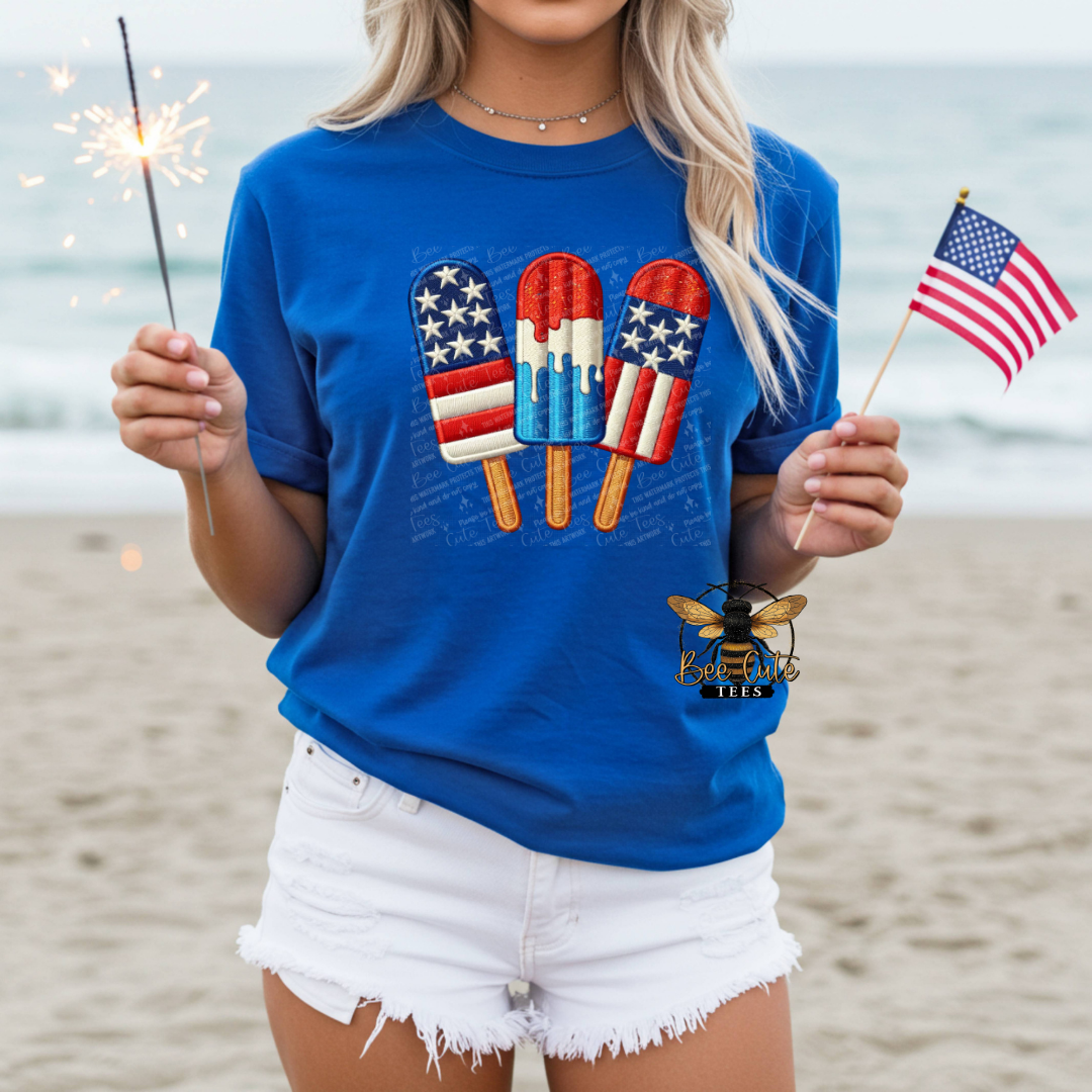 Person wearing a blue t-shirt with American flag-themed popsicles on a beach.
