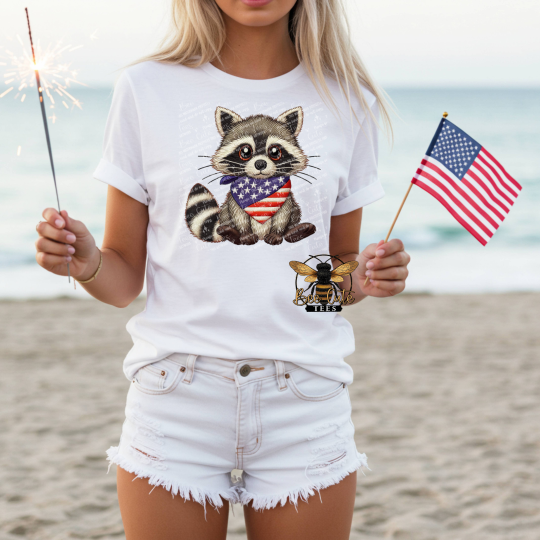 Person wearing a white t-shirt with a raccoon design holding an American flag on a beach.