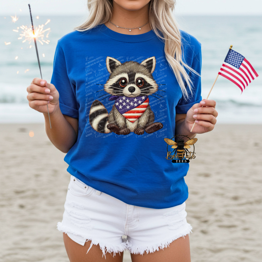 Person wearing a blue t-shirt with a raccoon design holding an American flag and sparkler on a beach.