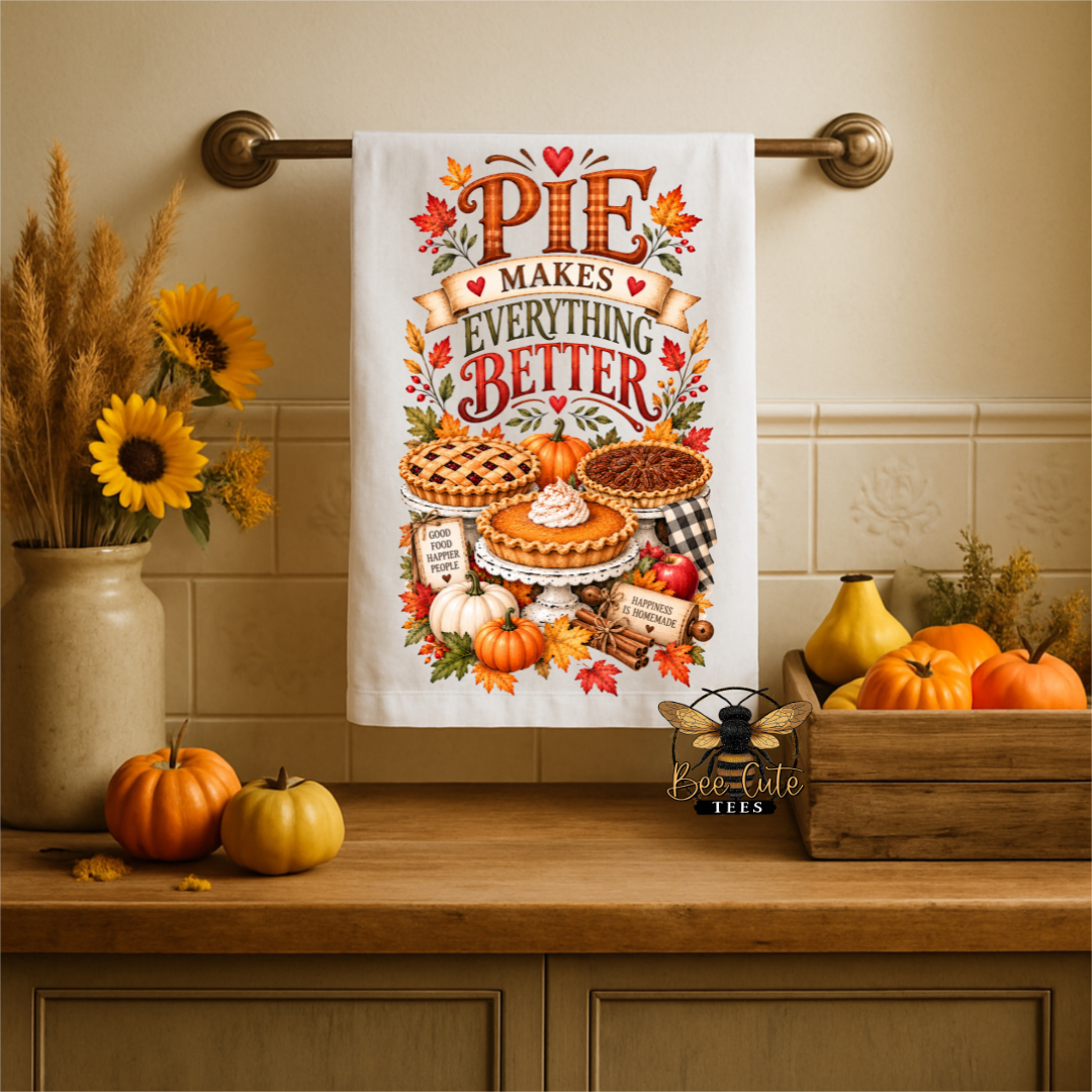 A white kitchen towel with a colorful autumn 'Pie Makes Everything Better' graphic hangs on a brass towel bar in a warm, decorated kitchen setting with pumpkins, sunflowers, and a wooden crate.