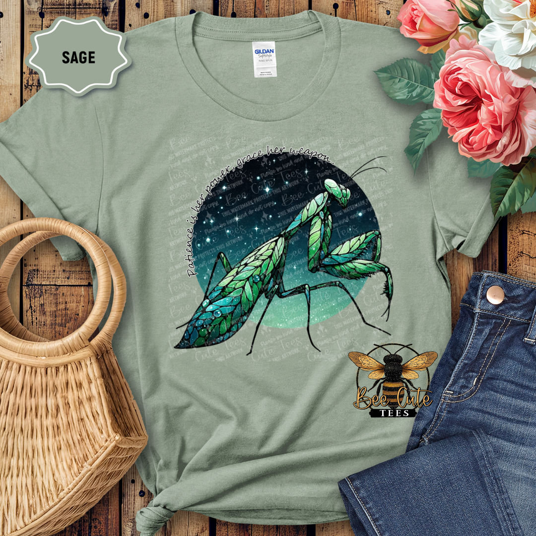 Sage green t-shirt with praying mantis design, placed on a wooden surface with flowers and jeans.