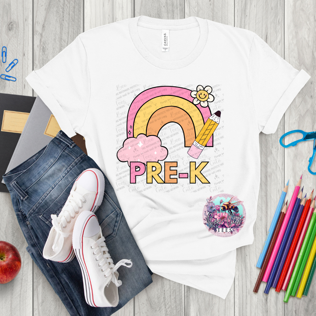 White t-shirt with a rainbow and 'PRE-K' design on a wooden surface with jeans, sneakers, and coloring supplies.