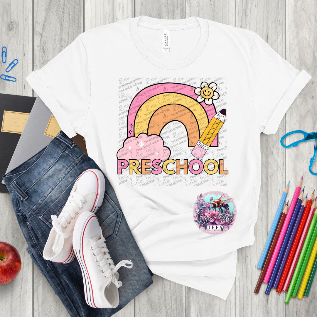 White t-shirt with 'PRESCHOOL' design on a wooden surface with jeans, shoes, and stationery items.