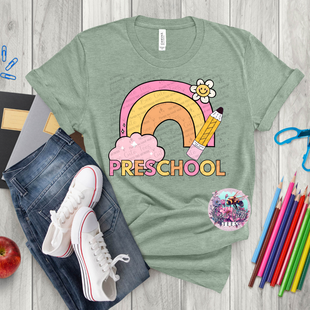 Green t-shirt with rainbow and pencil design labeled 'Preschool' on a wooden surface with school supplies.