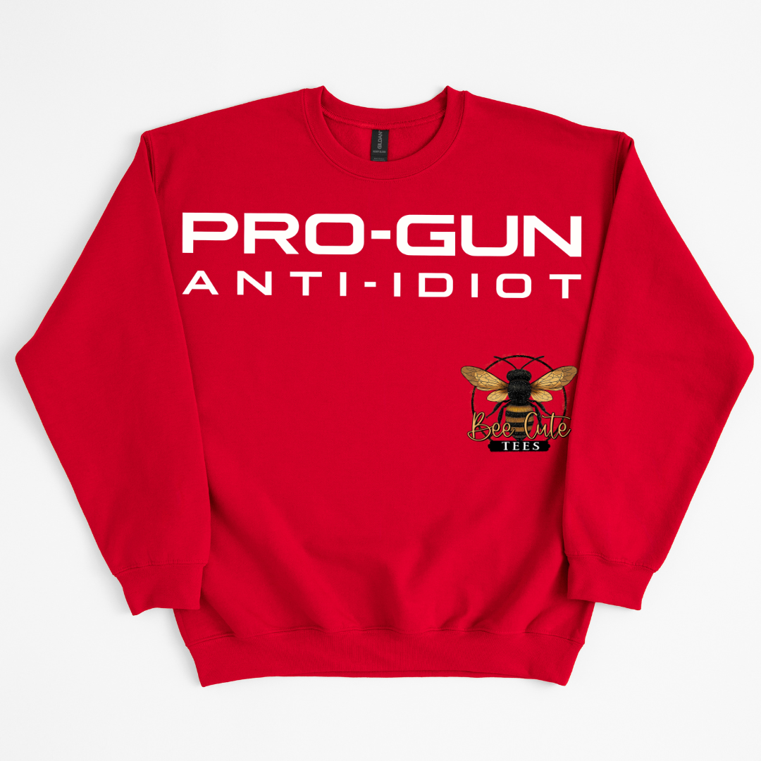 A red crewneck sweatshirt featuring the text 'PRO-GUN ANTI-IDIOT' in bold white letters across the chest, with a small bee logo printed on the lower right chest, displayed on a clean white background.