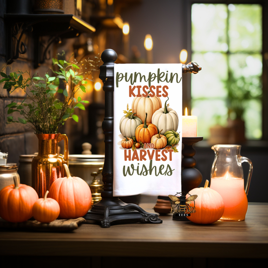 A decorative vertical tea towel with a white background and autumn illustration, featuring the text 'pumpkin KISSES and HARVEST wishes' with pumpkins in the center. The towel is displayed on a black stand on a wooden table surrounded by pumpkins, a copper vase with greenery, and warm ambient lights.