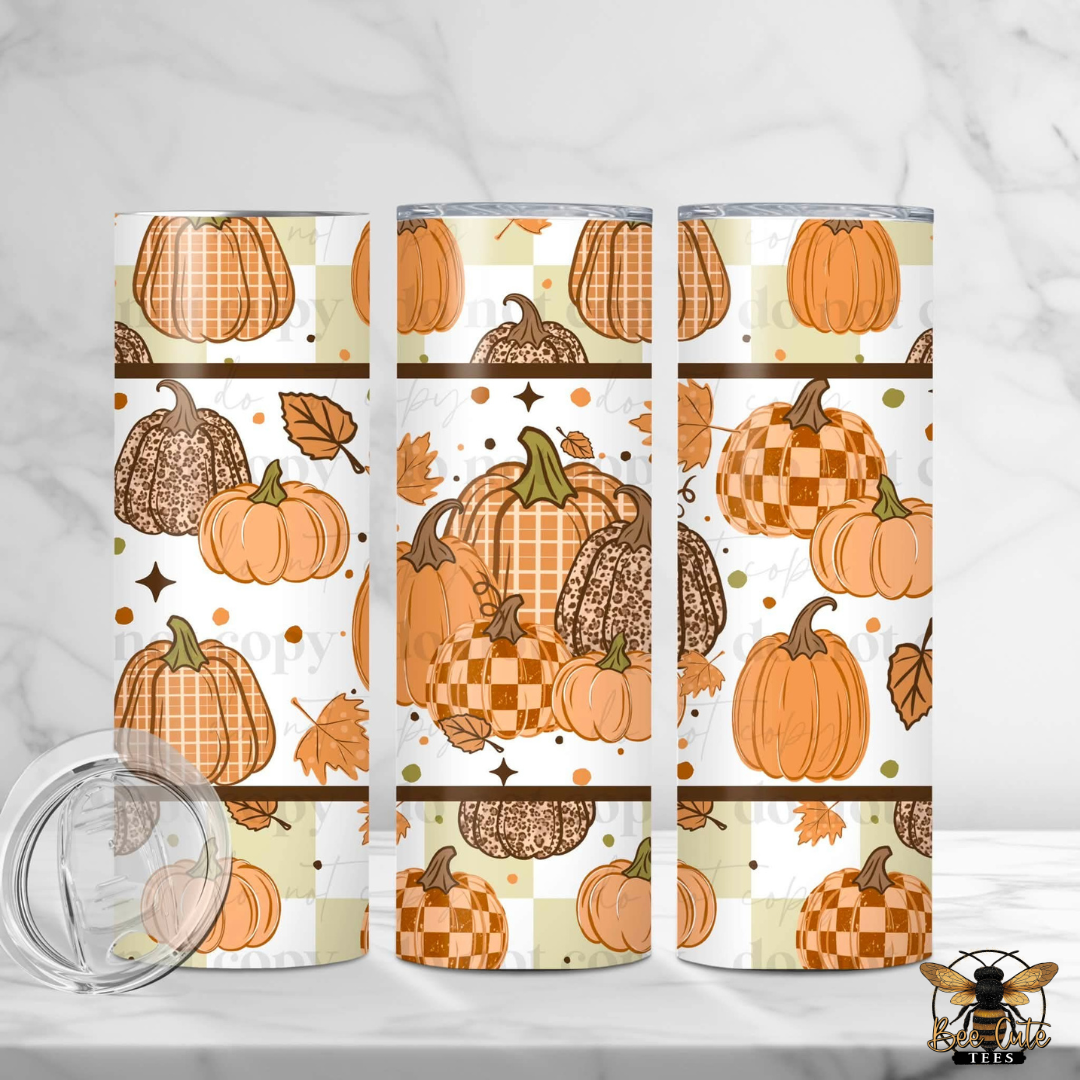 Three insulated tumblers with a fall pumpkin pattern are displayed on a white marble surface with a glass lid in the foreground. The pattern features orange and brown pumpkins, plaid and checkered motifs, and autumn leaves on a white background.