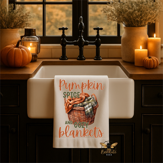 A white kitchen towel with a fall-themed graphic reading 'Pumpkin Spice and Cozy Blankets' hangs over a farmhouse sink. The scene is styled with pumpkins, candles, and dried plants in a warm autumn kitchen setting.