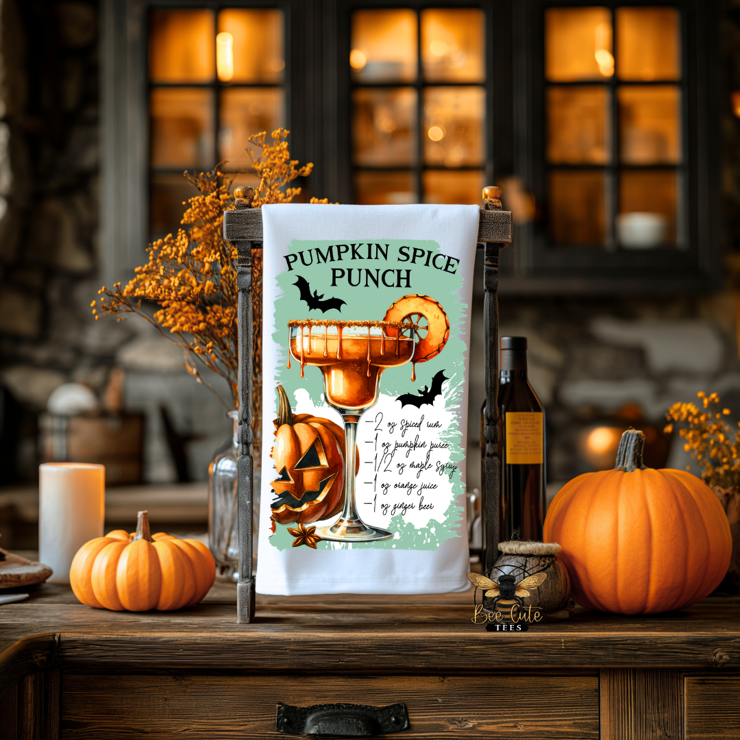 A white tea towel with a colorful 'Pumpkin Spice Punch' cocktail graphic and recipe print, displayed on a rustic wooden stand on a wooden table. The scene is decorated for Halloween with pumpkins, a candle, and a bottle of wine in a cozy, warmly lit indoor setting.