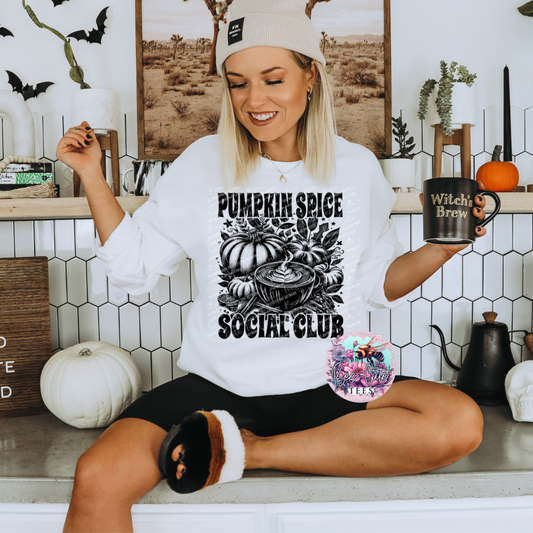 Woman wearing a 'Pumpkin Spice Social Club' shirt and holding a mug in a cozy room with Halloween decorations.