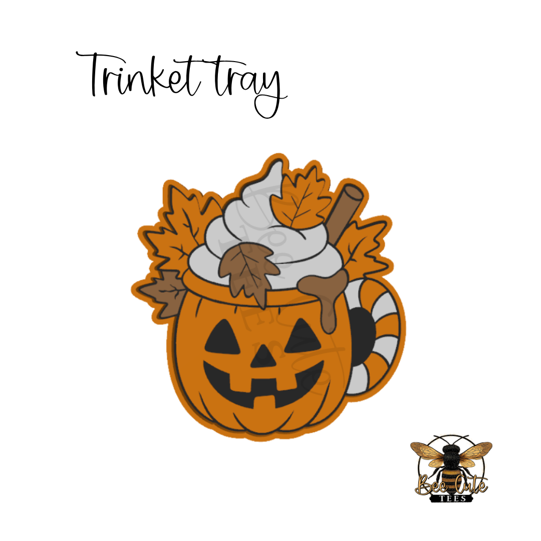 A Halloween-themed 'Trinket tray' design featuring a jack-o'-lantern mug filled with whipped cream, autumn leaves, and a cinnamon stick, shown on a white background with a bee logo in the bottom right.