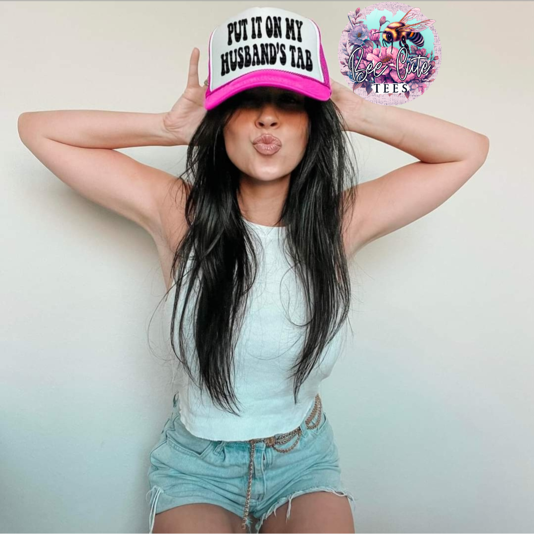 Woman wearing a white and hot pink trucker hat with “Put It On My Husband’s Tab” text, styled with a casual tank top and denim shorts, posing playfully against a neutral background.
