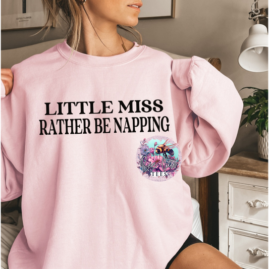 Person wearing a pink sweatshirt with 'Little Miss Rather Be Napping' text, sitting indoors.