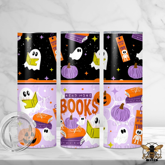 Three tall insulated tumblers with Halloween-themed wraparound prints are displayed on a light marble surface. The designs feature black, orange, purple, and white illustrations including ghosts reading books, pumpkins, stars, and the words 'READ MORE BOOKS' and 'Halloween TBR'.