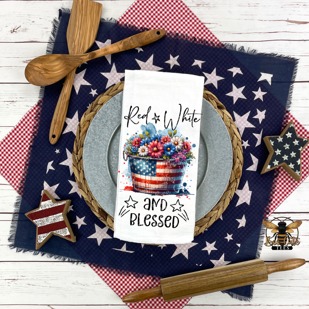 White kitchen towel displayed on a patriotic table setting, featuring a red, white, and blue floral arrangement in an American flag bucket with the phrase “Red White and Blessed,” surrounded by star-patterned linens, wooden utensils, and rustic decor.
