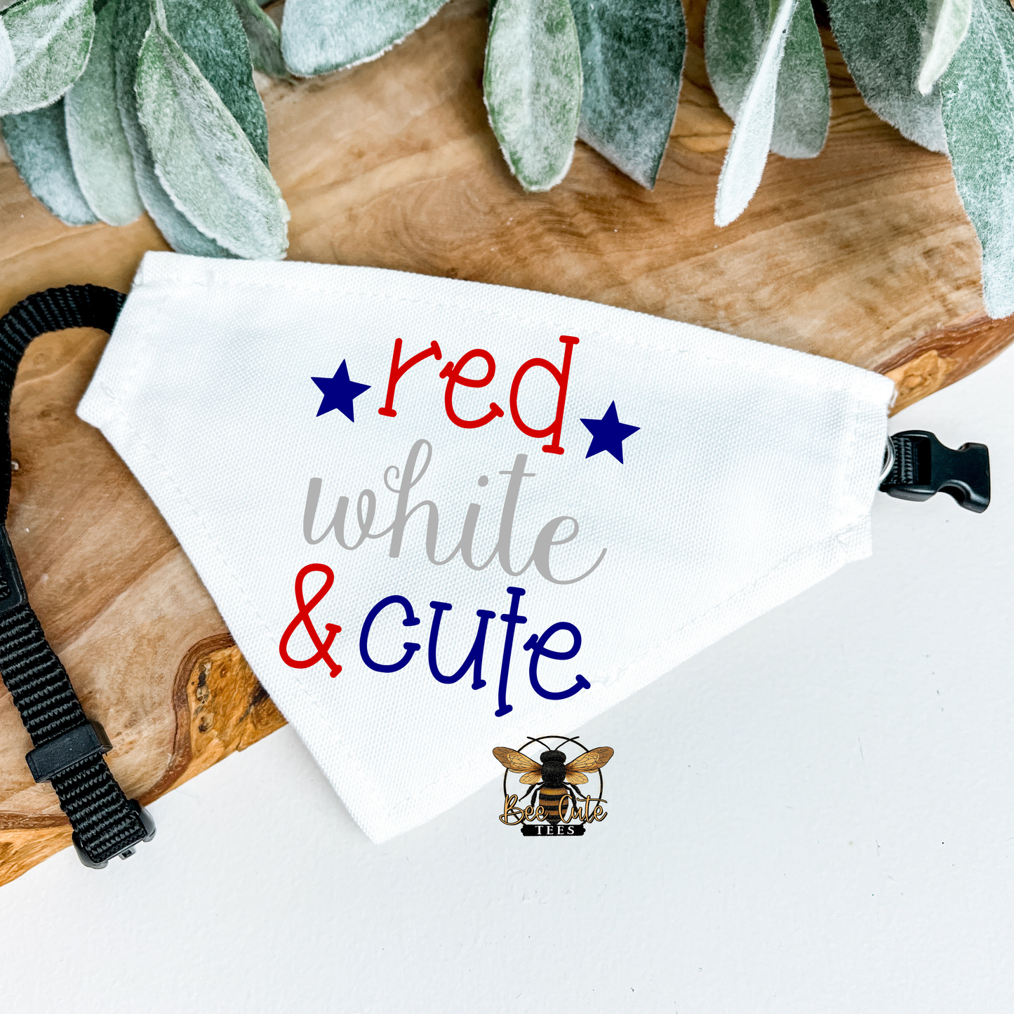 White bandana with 'red white & cute' text and bee logo on a wooden surface with greenery.