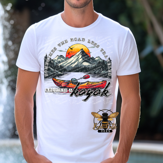 Man wearing a white graphic tee featuring colorful kayaks on a lake with mountain scenery and a sunset background, paired with the phrase “Sometimes the Road Less Traveled Requires a Kayak.”
