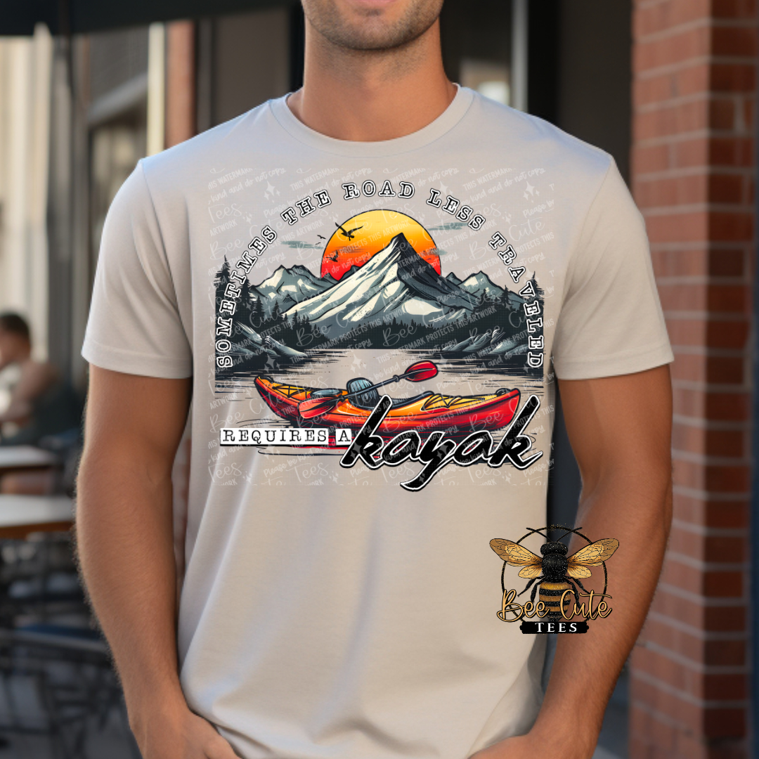 Man wearing a beige graphic tee featuring colorful kayaks on a lake with mountain scenery and a sunset background, paired with the phrase “Sometimes the Road Less Traveled Requires a Kayak.”
